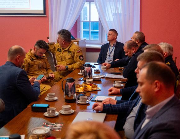 Grupa około dziesięciu mężczyzn w garniturach i mundurach siedzi przy dużym, drewnianym stole konferencyjnym w pomieszczeniu o czerwonych ścianach. Piją kawę i omawiają dokumenty.