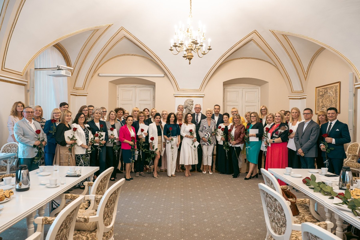 grupa około kilkudziesięciu osób kobiet i mężczyzn pozuje do zdjęcia w zabytkowej reprezentacyjnej, jasnej sali, są ubrani elegancko, uśmiechnięci, niektórzy trzymają kwiaty