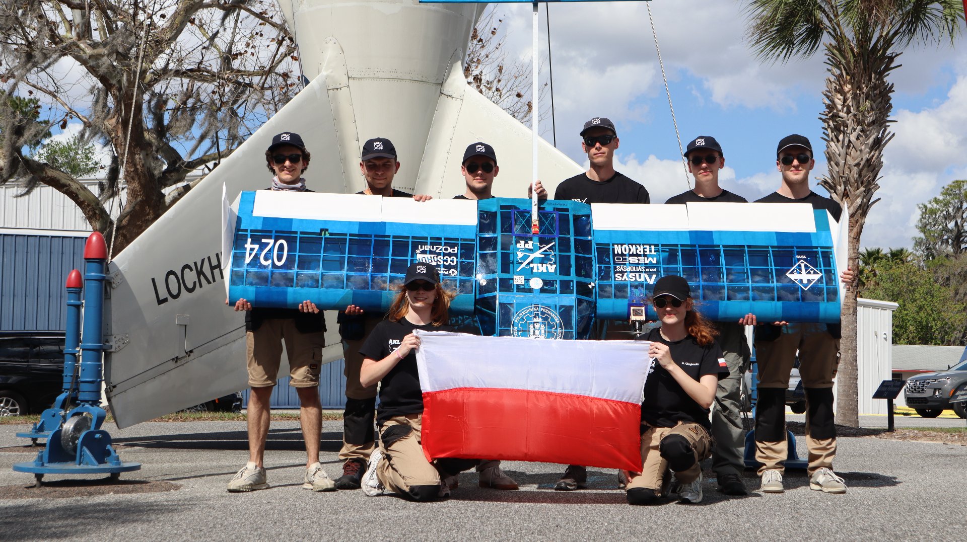Grupa ośmiu osób pozuje na zewnątrz przed ogonem dużego, jasnego samolotu z napisem "LOCKH". Sześciu mężczyzn stoi w rzędzie, trzymając szeroki, błękitny model samolotu z logo Politechniki Poznańskiej i numerem "024". Przed nimi klęczą dwie kobiety, trzymając biało-czerwoną flagę. Wszyscy członkowie zespołu ubrani są w czarne koszulki i czarne czapki z daszkiem. Po prawej stronie widoczna jest palma, a w tle błękitne niebo z chmurami.
