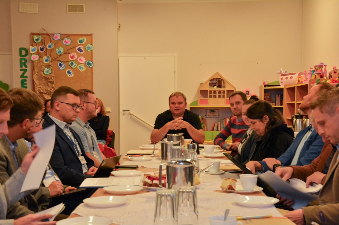 grupa osób kobiet i mężczyzn siedzi przy podłużnym stole, prowadzą spotkanie, na stole dokumenty, laptopy, termosy do napojów, taleczyki i filiżanki