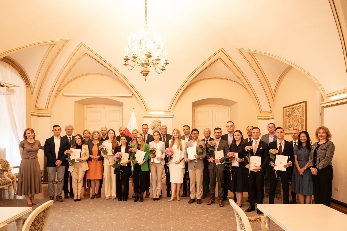grupa osób kobiet i mężczyzn w różnym wieku pozuje do wspólnego zdjęcia, są elegancko ubrani, niektórzy z nich trzymają kwiaty, znajdują się w eleganckiej, zabytkowej sali z żyrandolem