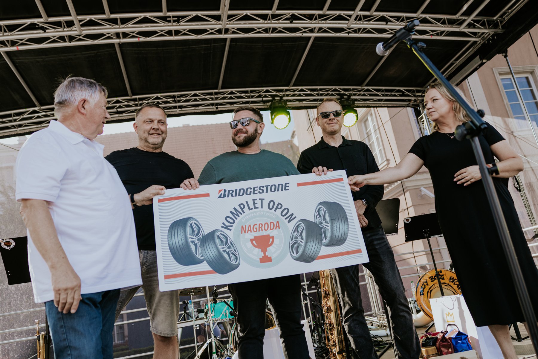 Grupa osób na scenie podczas uroczystego wręczenia pamiątkowej tablicy symbolizującej nagrodę w postaci kompletu opon Bridgestone.