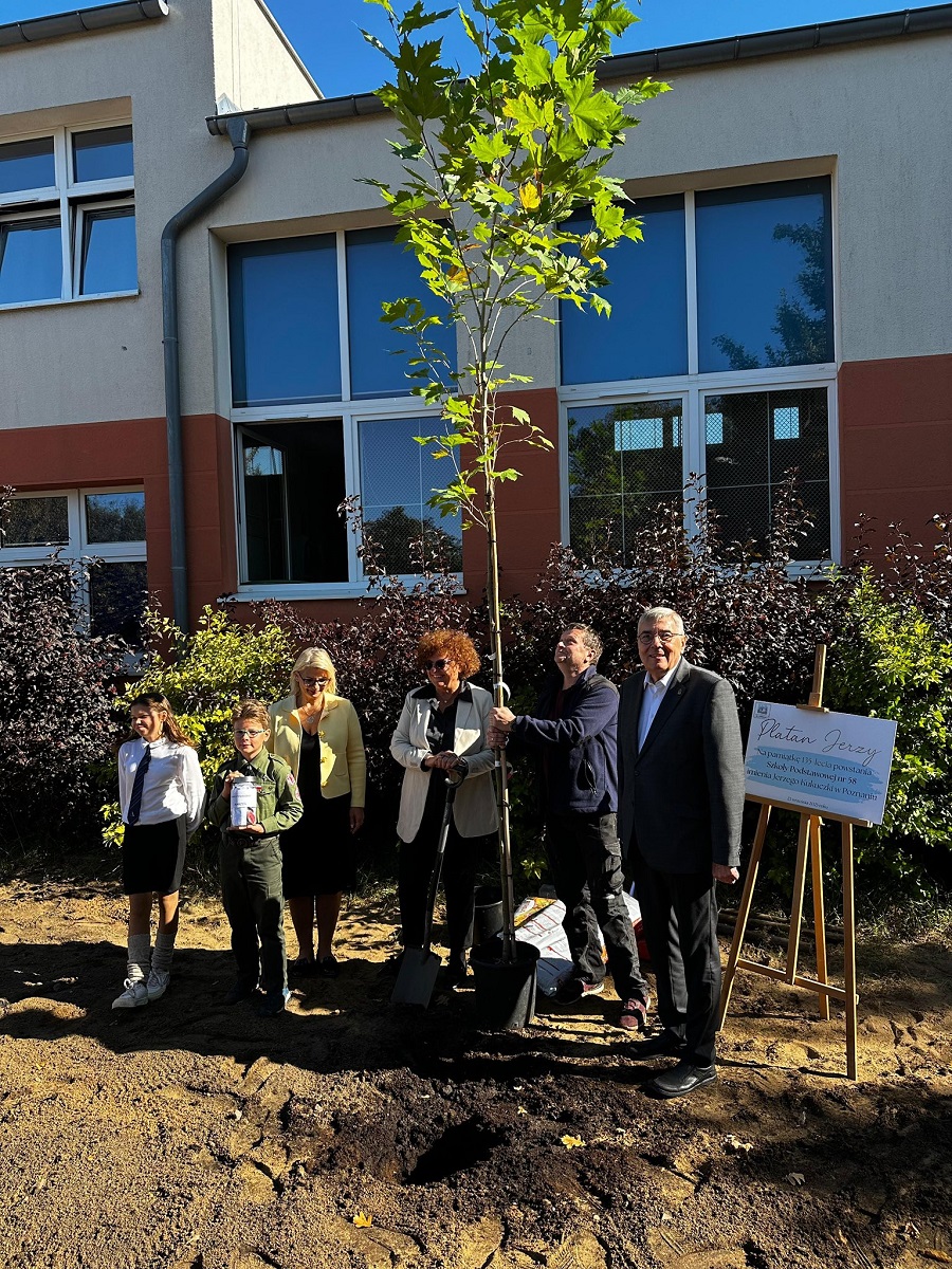 grupa osób pozuje do zdjęcia przy nowo posadzonym drzewie przed budynkiem szkolnym