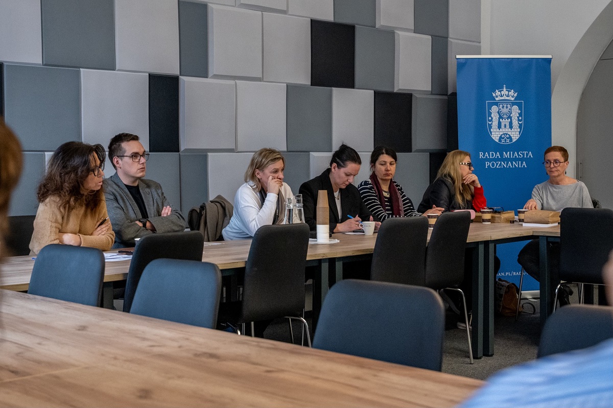 grupa osób siedzi za stołem w sali konferencyjnej, w tle baner z herbem Poznania i napisem RADA MIASTA POZNANIA - więcej w treści artykułu