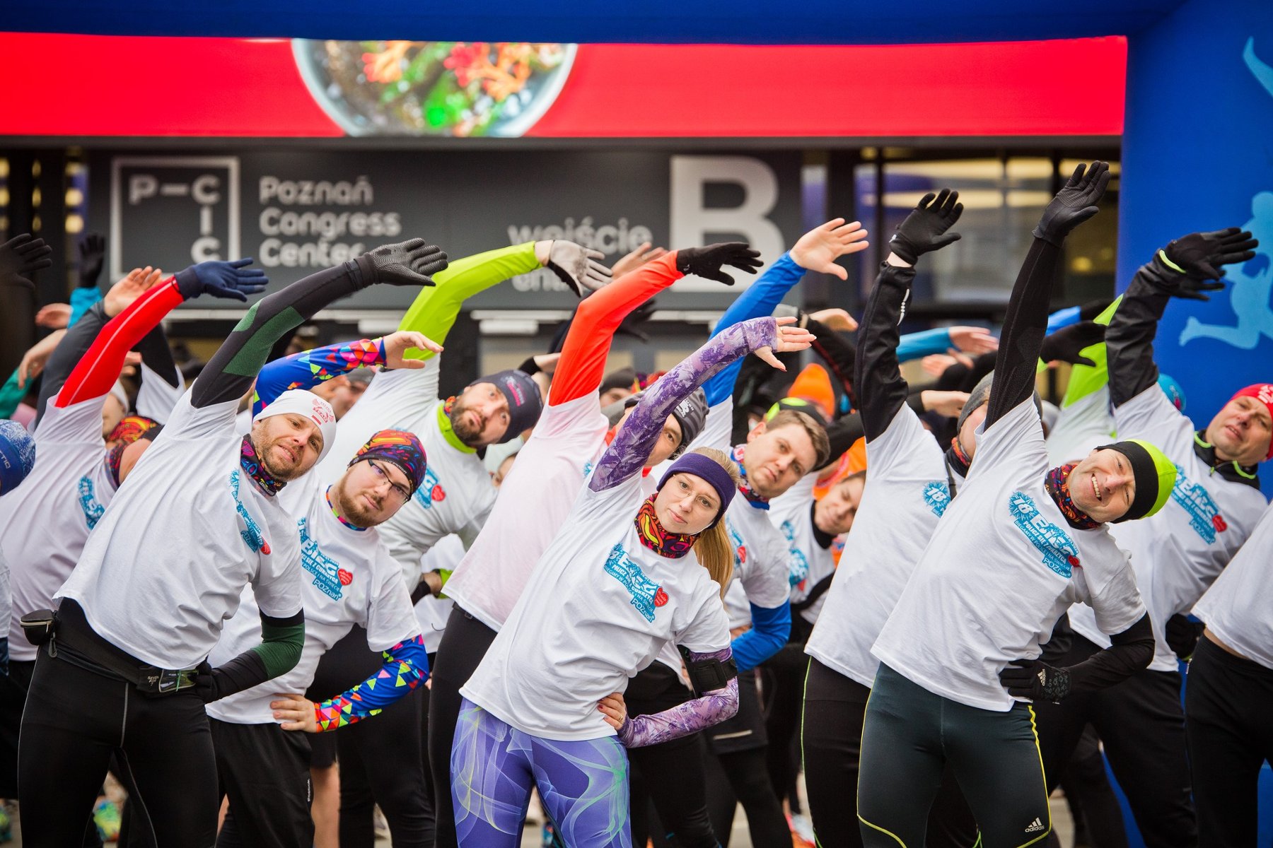 Grupa osób ubrana na sportowo z białymi koszulkami biegu rozciąga się przed biegiem charytatywnym. W tle start biegu i budynek.