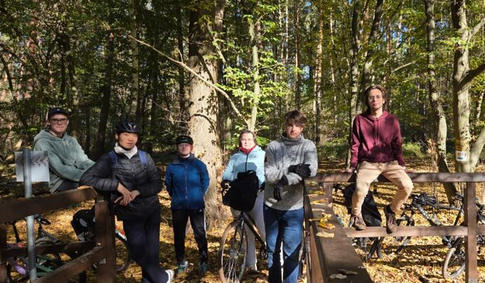 Grupa osób w jesiennym lesie, część osób na na sobie kaski rowerowe. Obok ludzi stoją rowery.