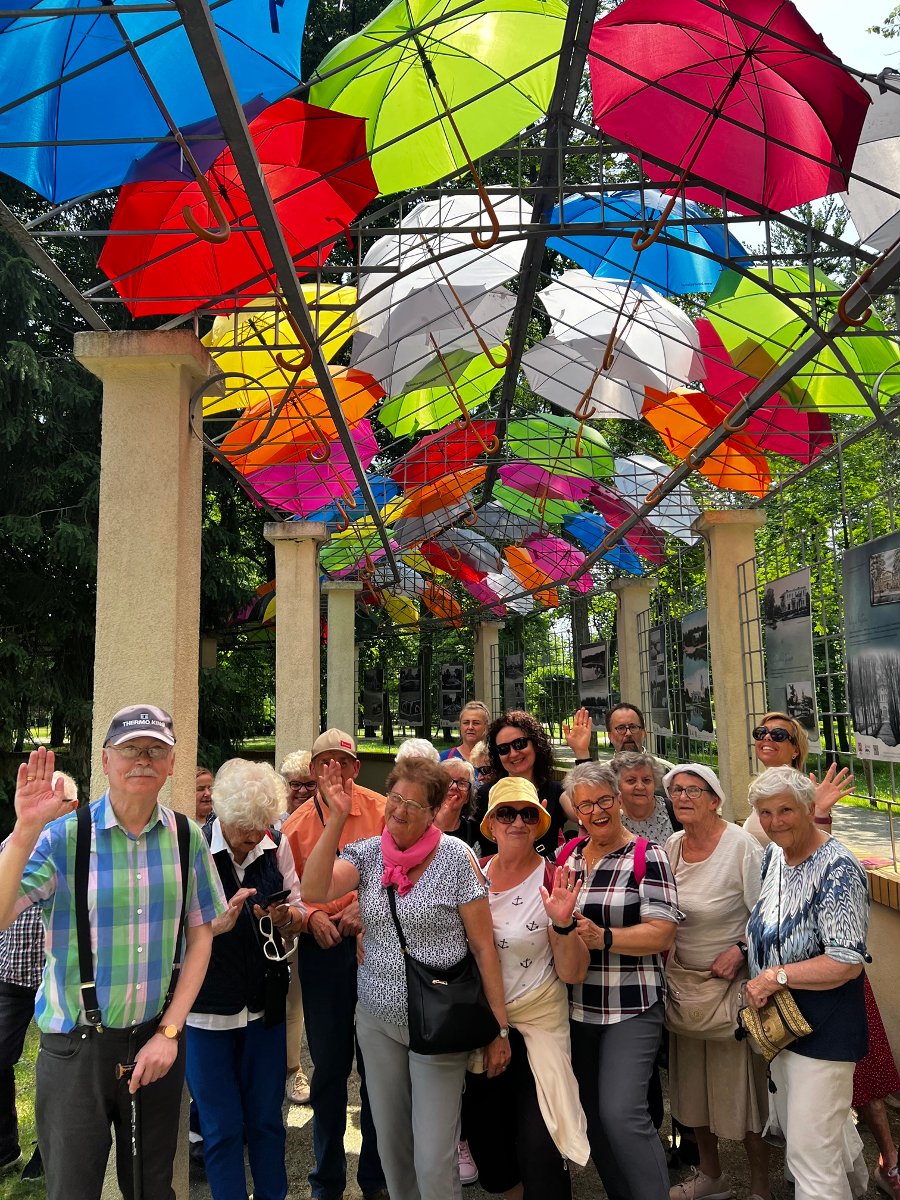 Grupa seniorów pod pergolą ozdobioną kolorowymi parasolami.