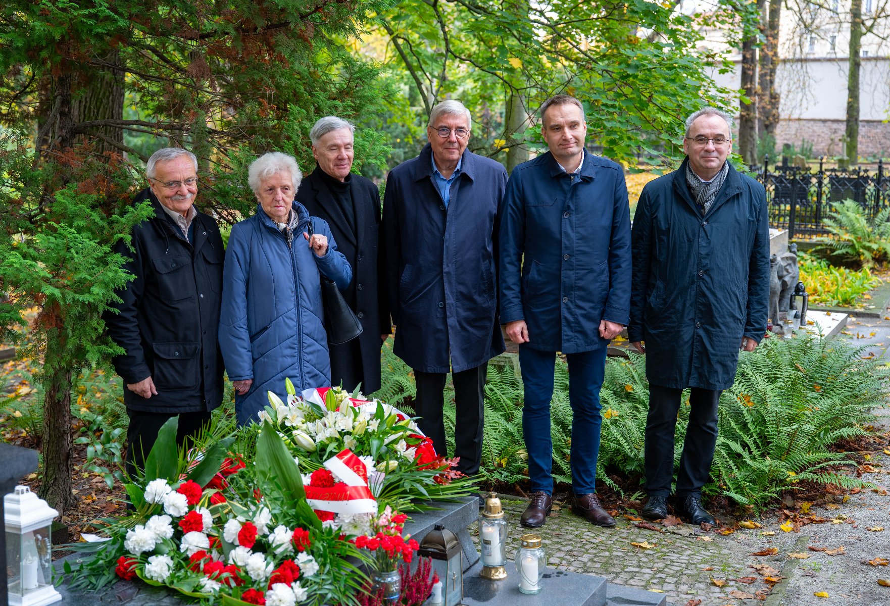Grupa sześciu osób, pięciu mężczyzn i jedna kobieta, w ciemnych okryciach wierzchnich, stoi na cmentarzu. Przed nimi grób z wieńcem z biało-czerwonych kwiatów, dwoma zniczami i lampionem. W tle widoczna zieleń drzew i nagrobki.