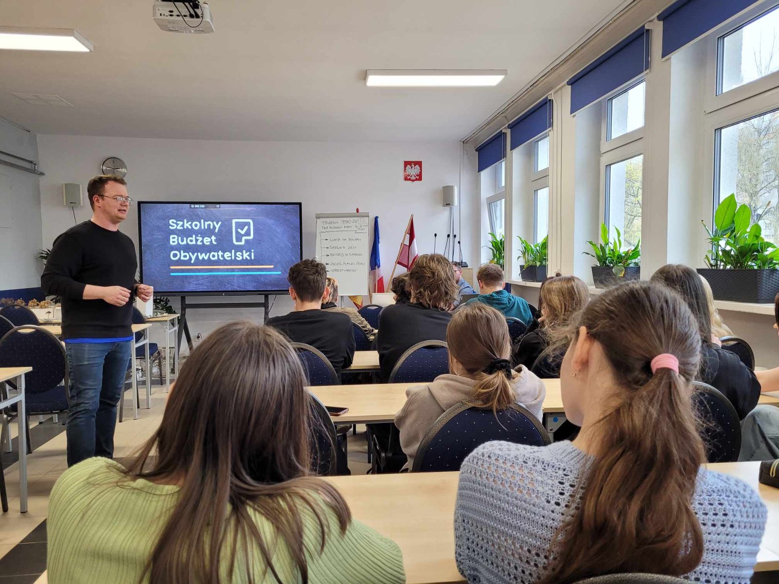 Grupa uczniów tyłem wraz z prowadzącym w klasie, w tle ekran z napisem "Szkolny Budżet Obywatelski"