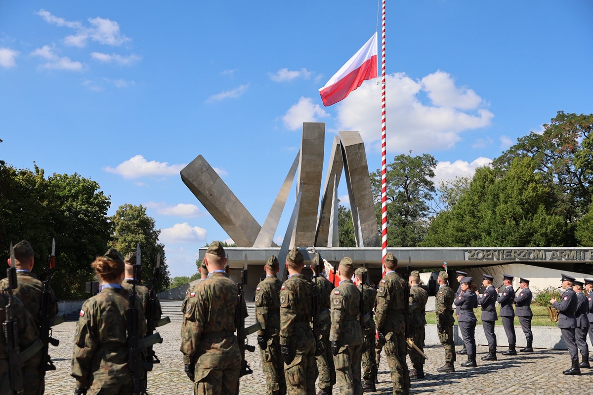 grupa żołnierzy stoi na placu przed pomnikiem, jest maszt a na nim flaga biało czerwona