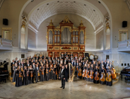 Grupowe zdjęcie orkiestry i dyrygenta. Muzycy trzymają w rękach instrumenty i pozują do fotografii.