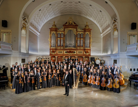 Grupowe zdjęcie orkiestry i dyrygenta. Muzycy trzymają w rękach instrumenty i pozują do fotografii.