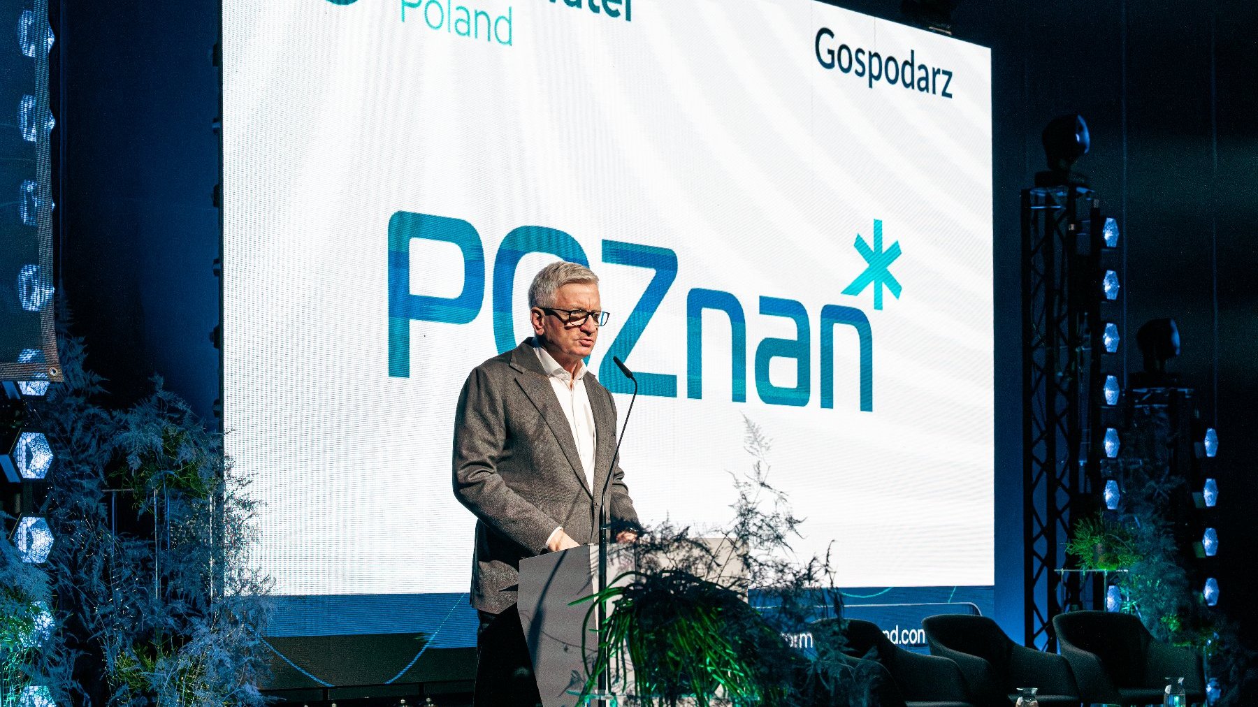 Ilustracja przedstawia Prezydenta Miasta Poznania Jacka Jaśkowiaka przemawiającego na tle planszy z napisem "Poznań" oraz "Gospodarz".