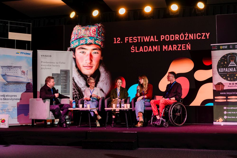 Jasnowłosa kobieta w okularach uśmiecha się szeroko do obiektywu, za nią baner festiwalowy.