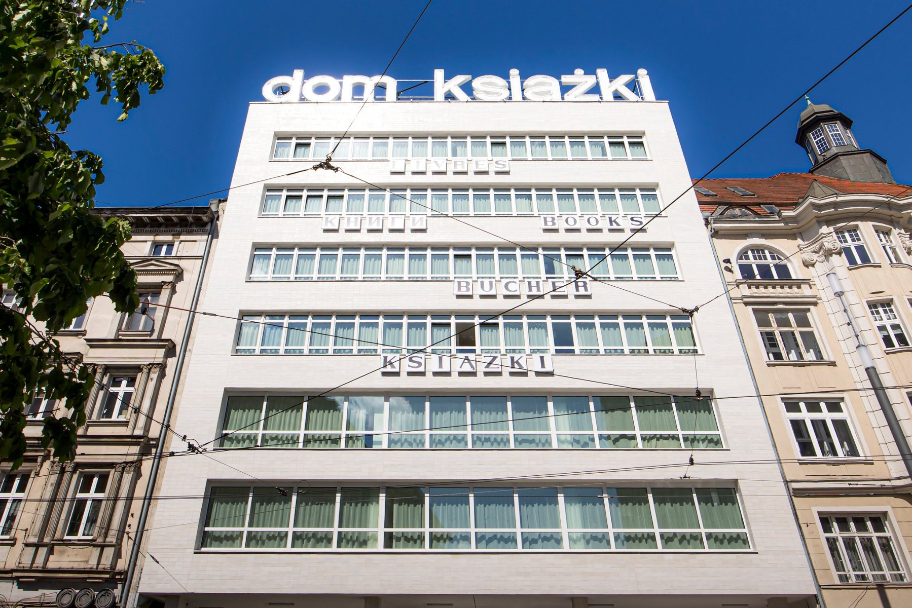 Jasny, kilkupiętrowy budynek modernistyczny z dużym napisem "dom książki" na dachu. Na elewacji, między rzędami okien, umieszczono napisy "książki" w różnych językach. Budynek sąsiaduje ze zdobionymi kamienicami, a przed nim widoczne są przewody trakcji tramwajowej na tle czystego, błękitnego nieba.