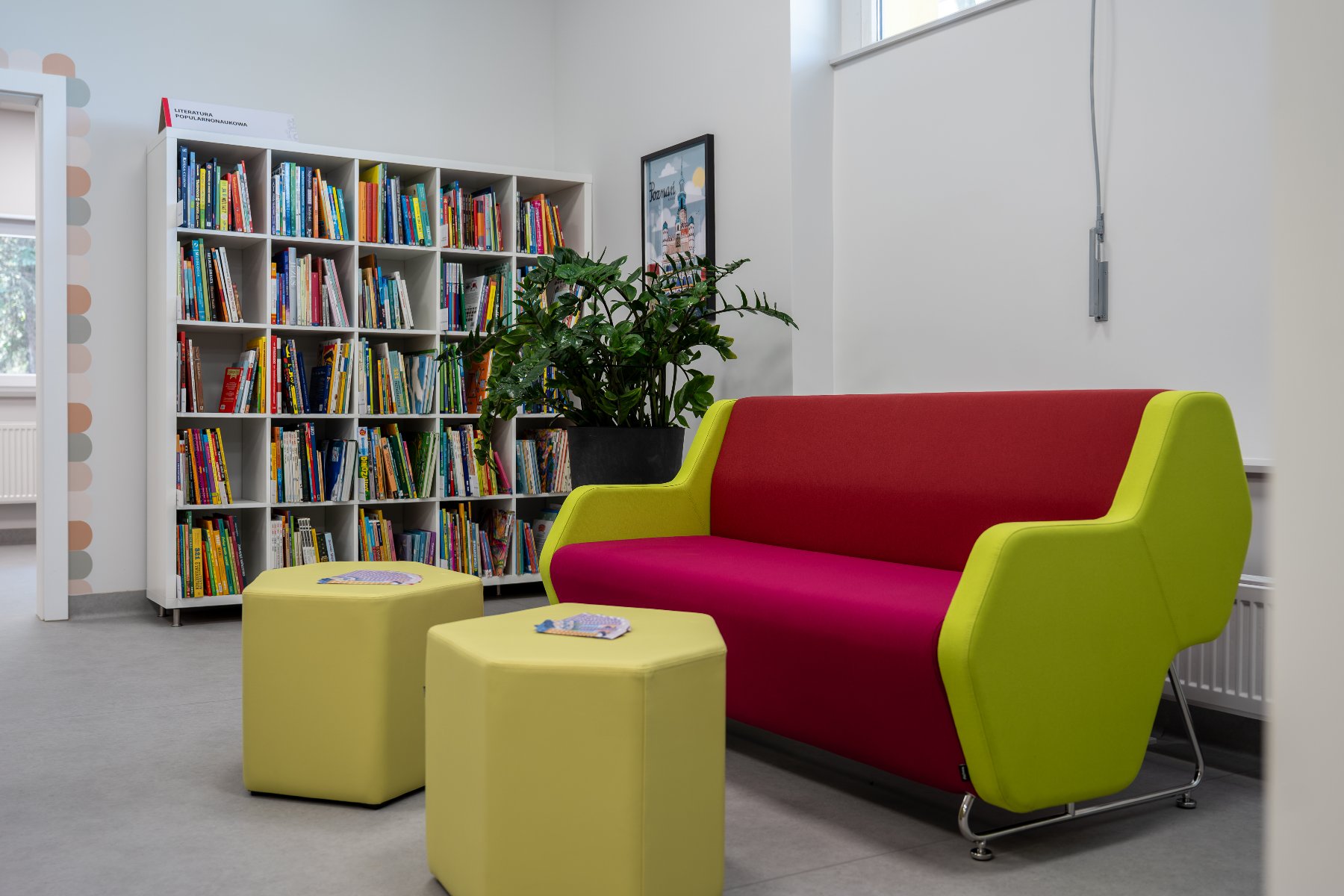 Kącik czytelniczy w bibliotece z dwukolorową, czerwono-zieloną kanapą, dwoma pufami, rośliną doniczkową oraz regałem pełnym książek w tle.