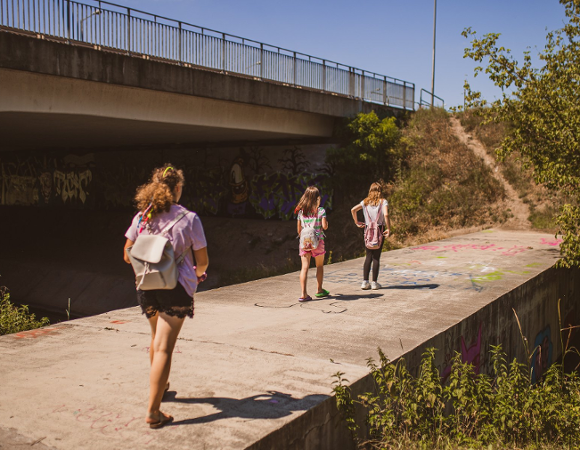 Kadr od tyłu przedstawia trzy dziewczynki idące betonową ścieżką obok wiaduktu drogowego. Pierwsza z lewej ma na sobie lawendową koszulkę, czarne koronkowe szorty i jasnoszary plecak; pozostałe dwie mają kolorowe plecaki i krótkie spodnie. Ściany pod wiaduktem pokryte są kolorowym graffiti. Obok ścieżki wznosi się porośnięte trawą i krzewami wzgórze, na które prowadzi wąska ścieżka. Nad wszystkim góruje niebieskie, bezchmurne niebo, wskazując na słoneczny dzień.