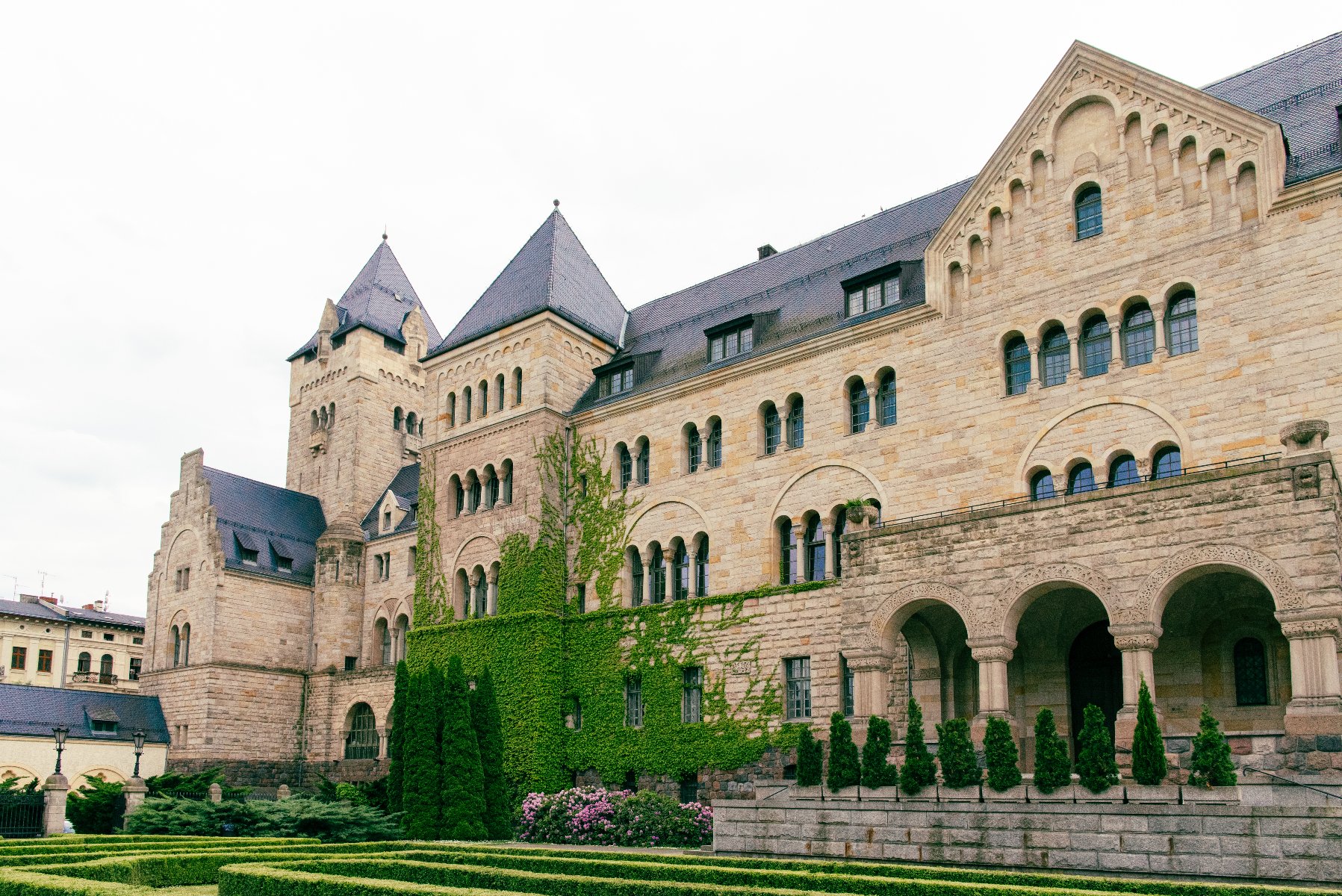 Kamienna konstrukcja z wieloma elementami architektonicznymi, takimi jak łukowate okna, spiczaste dachy i wieże. Front budynku zawiera wiele rzędów okien, niektóre grupowane po trzy lub więcej. Wyraźne wejście łukowe, wspierane przez kolumny. Otoczenie budynku obejmuje zadbaną zieleń: żywopłoty, drzewa i kwitnące rośliny.