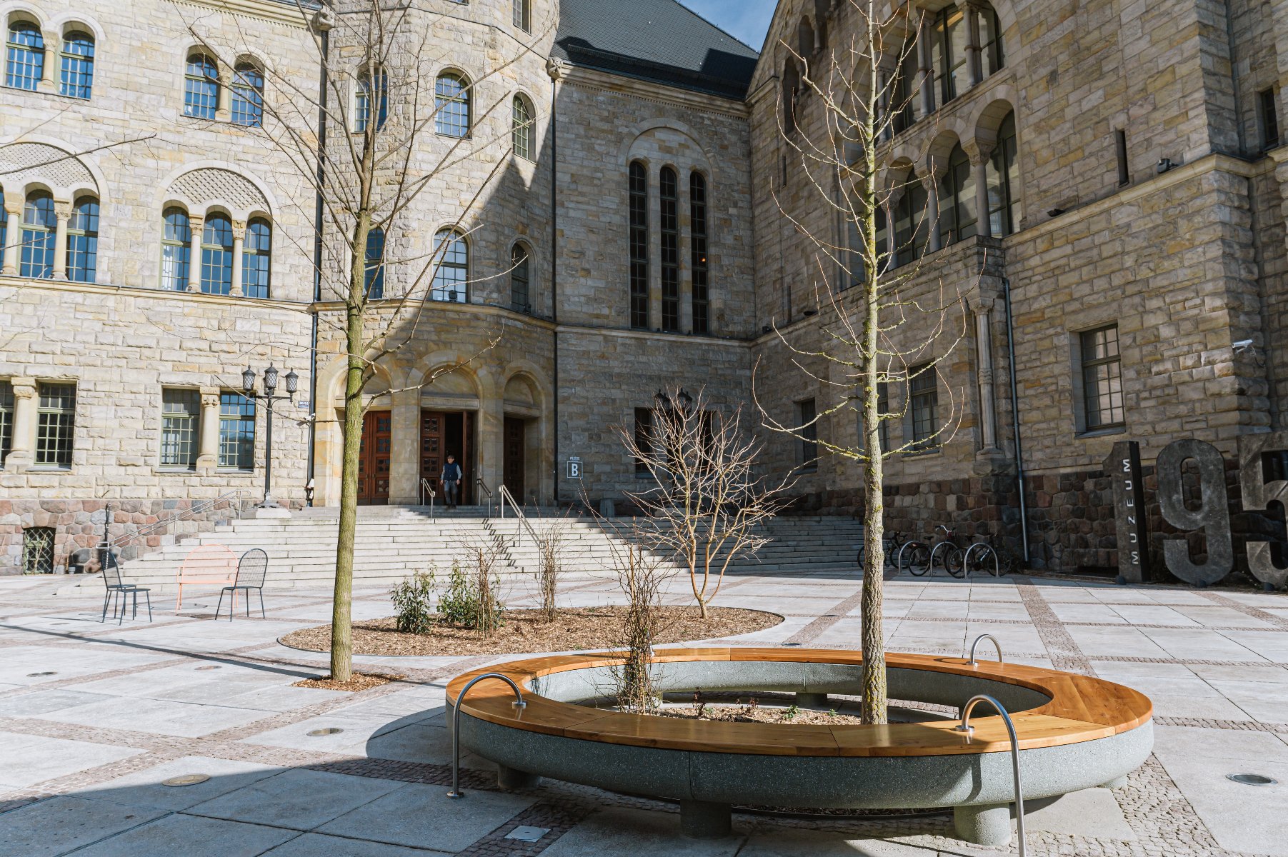 Kamienny budynek z wieloma łukowatymi oknami oraz centralnym wejściem z prowadzącymi do niego schodami. Na pierwszym planie okrągła ławka wokół drzewa, a w pobliżu kolejne drzewo i kilka mniejszych roślin.