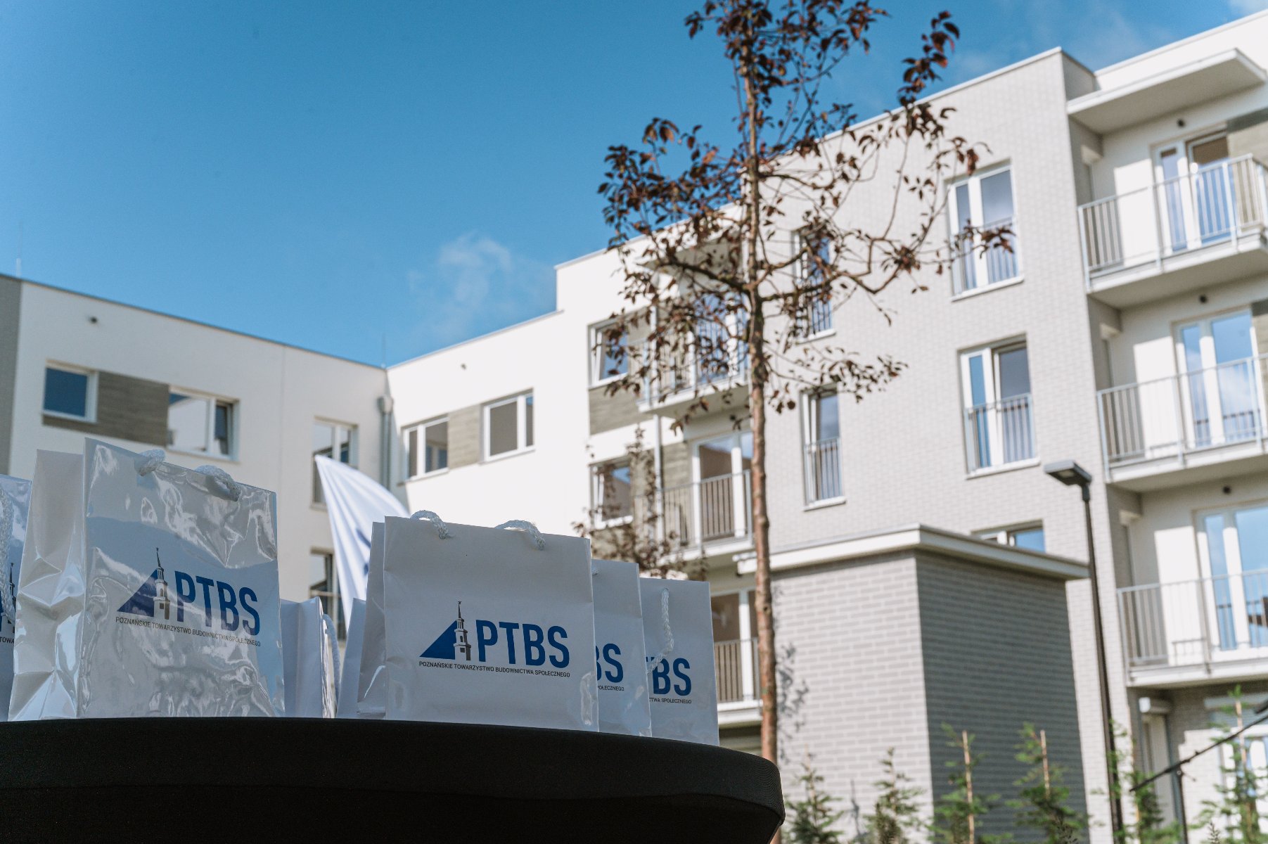 Kilka białych toreb z logo "PTBS' ustawionych na czarnym stole. W tle nowoczesny budynek mieszkalny z wieloma piętrami, balkonami i oknami. Widać również drzewo o rzadkich liściach oraz jasne niebo z kilkoma chmurami. Sceneria sugeruje wydarzenie związane z działalnością deweloperską lub prezentacją projektu mieszkaniowego.