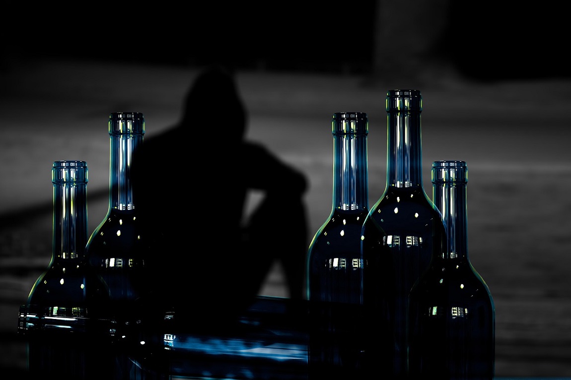 kilka butelek, za nimi ludzka postać w kapturze, odwrócona w pozycji siedzącej, zdjęcie jest zaciemnione