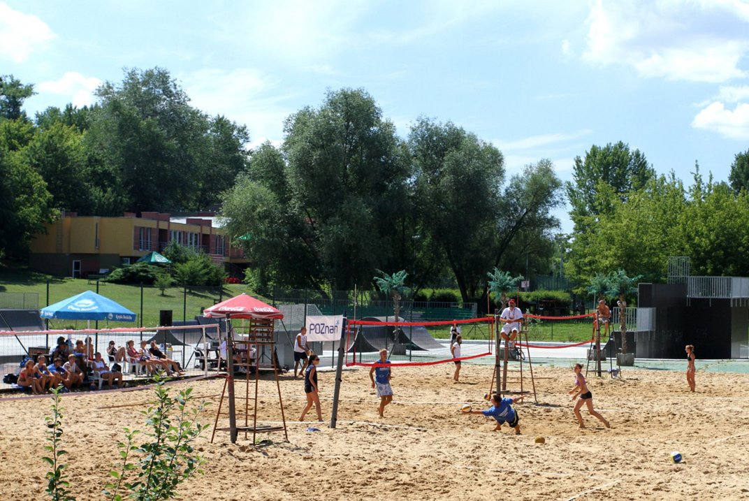 kilka osób gra w siatkówkę plażową, inne osoby oglądają mecz, jest słoneczna pogoda, za boiskiem plażowym widać skatepark i dalej drzewa