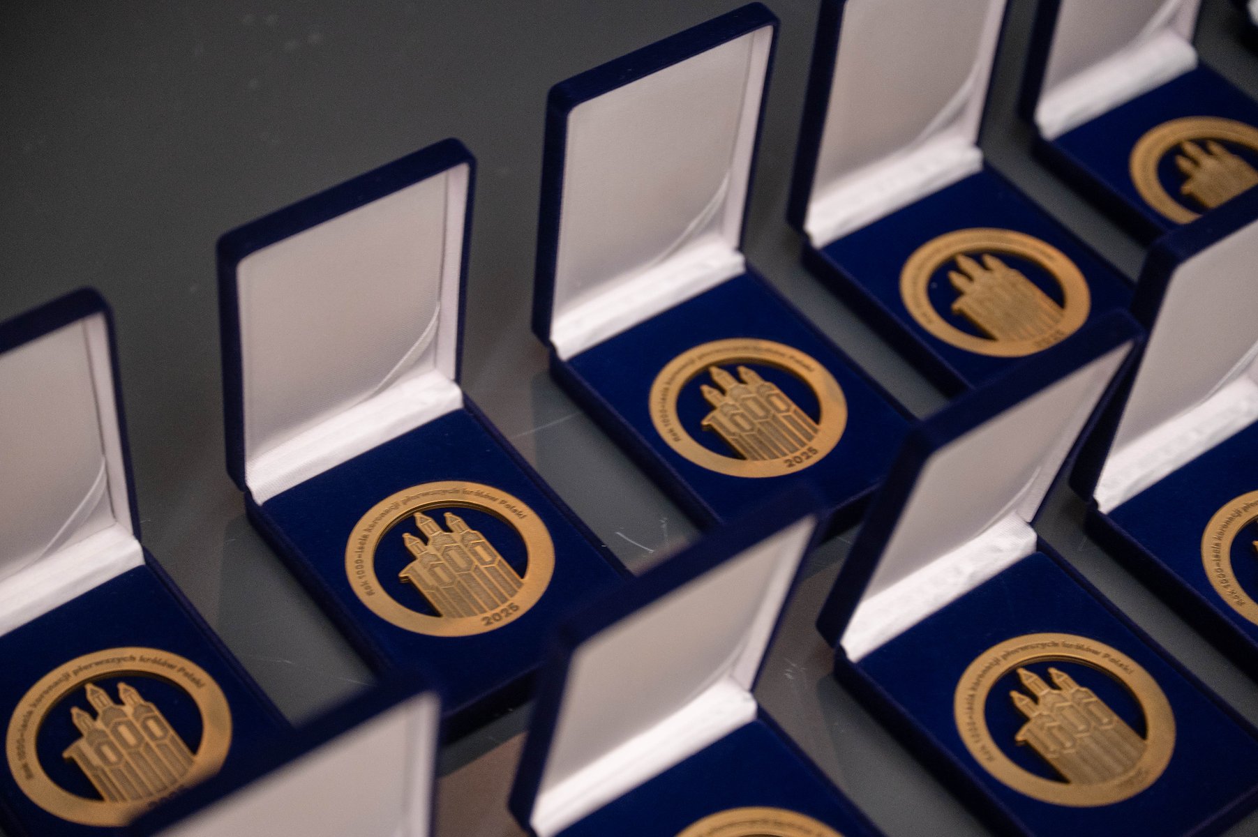 Kilka otwartych granatowych pudełek na aksamitnej wyściółce. W każdym pudełku znajduje się okrągły, złoty medal z symbolami Poznania oraz dużym napisem i datą w cudzysłowie: "1000 LAT" i "2025".