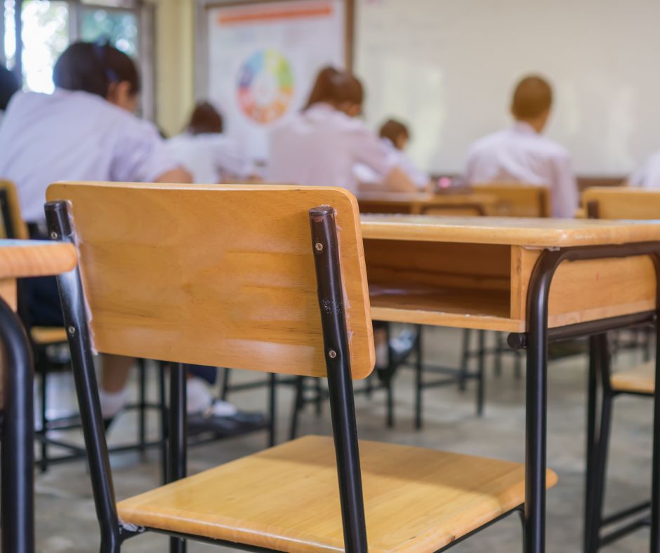 klasa szkolna, na pierwszym planie puste drewniane krzesło i stolik, na dalszym planie młodzi ludzie siedzący w ławkach i uczestniczący w zajęciach.