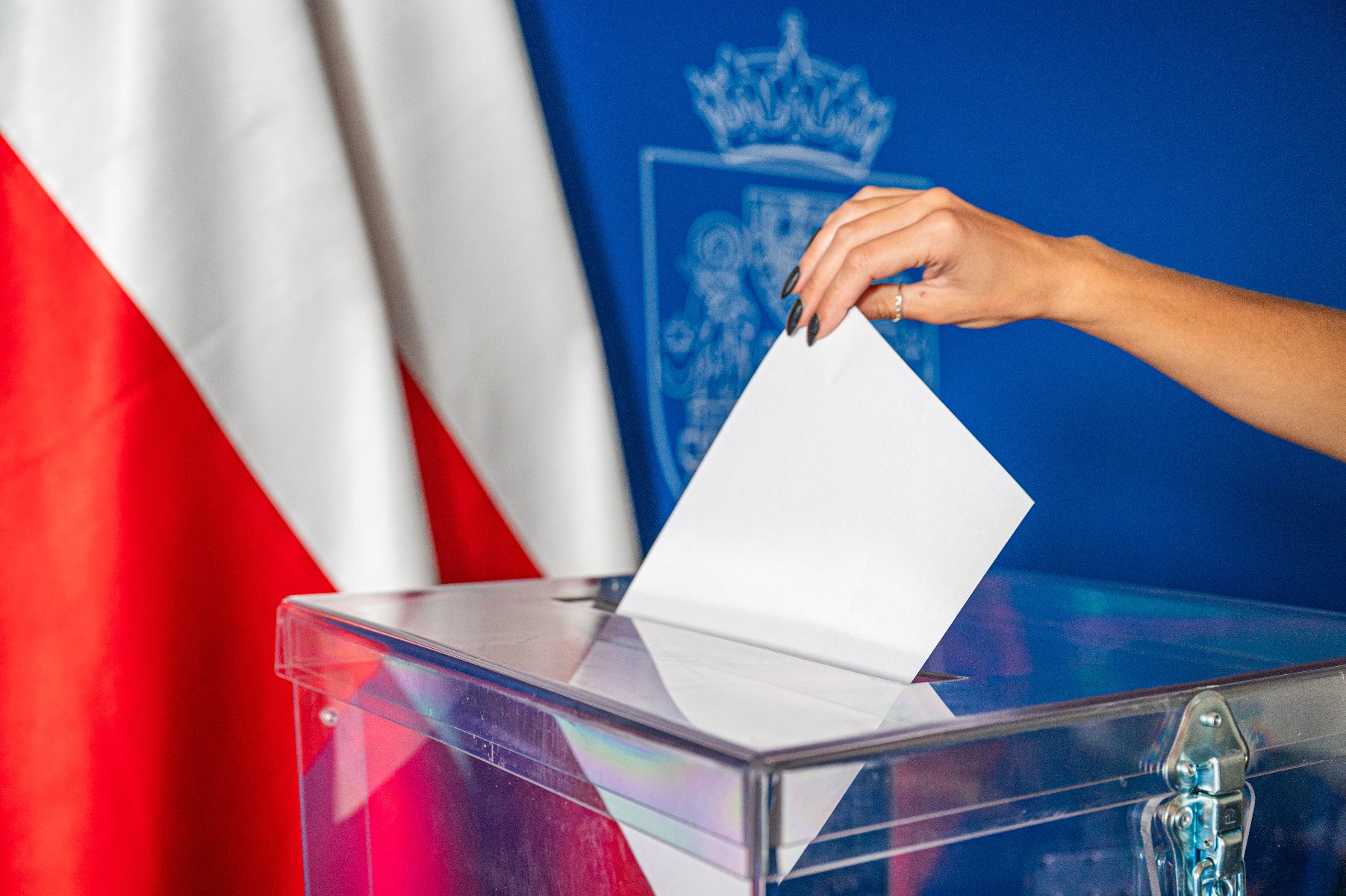 Kobieca ręka wrzuca kartkę do urny. W tle biało-czerwone flagi i herb Poznania na niebiskeim tle