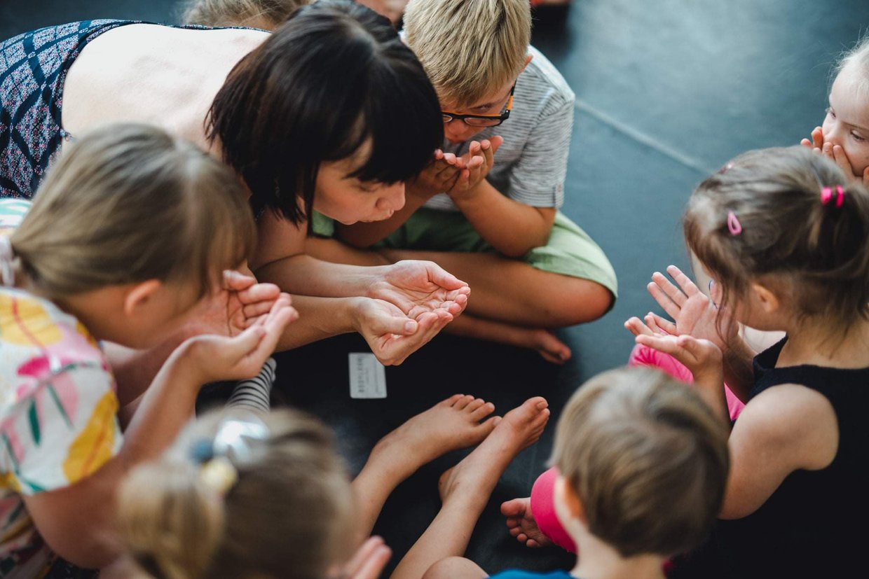 Kobieta i grupa małych dzieci siedzą na podłodze blisko siebie. Nachylają się do siebie i chuchają w swoje dłonie.