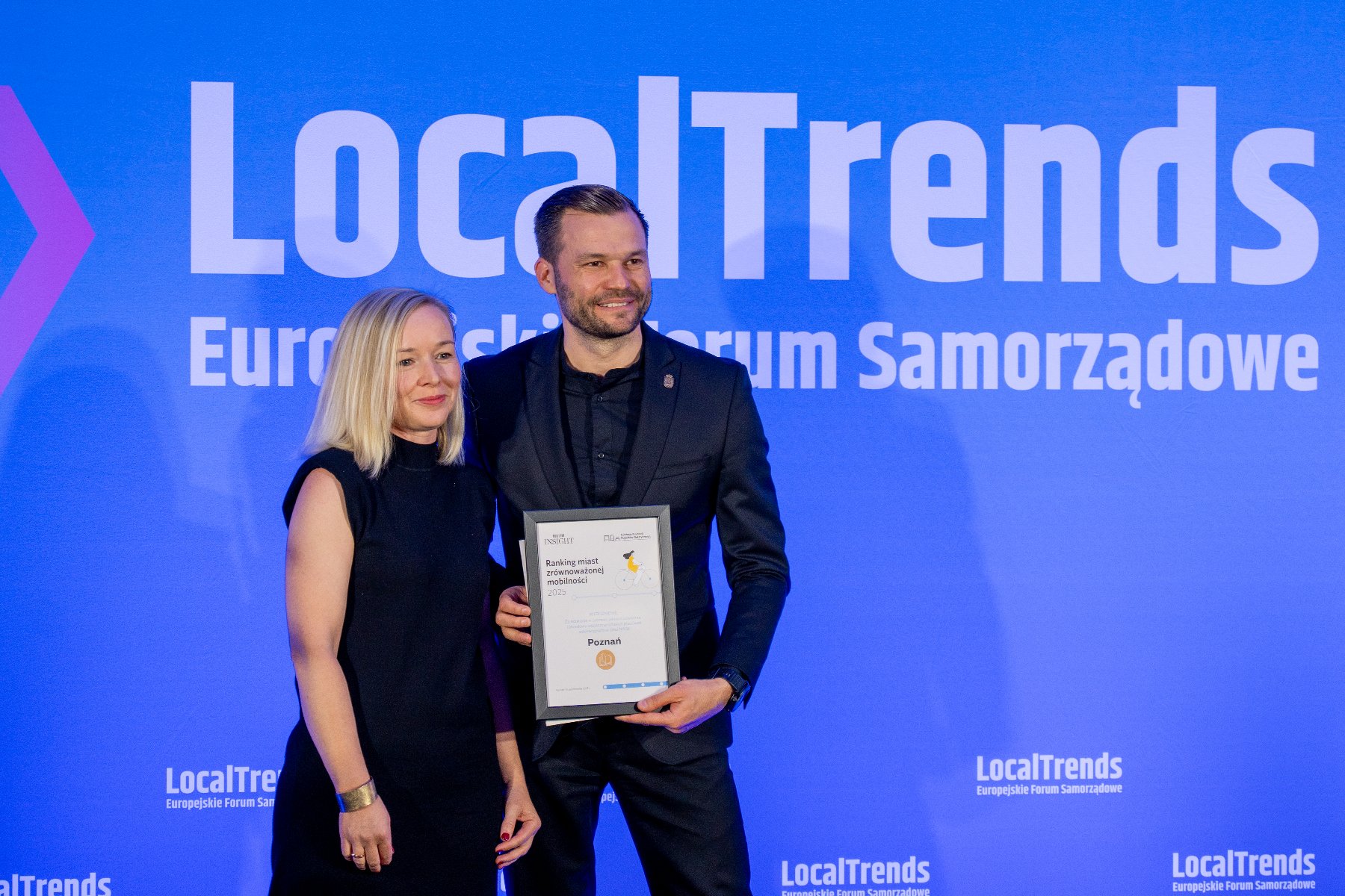 Kobieta i mężczyzna stoją na tle niebieskiego ekranu z logiem LocalTrends. Mężczyzna trzyma w rękach dyplom z napisem: "Poznań". Oboje uśmiechają się.