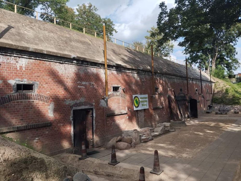 Kobieta trzyma za rękę dziecko, wspólnie wchodzą do wnętrza wąskiego, ciemnego korytarza w forcie, lekko tylko oświetlonego latarką.
