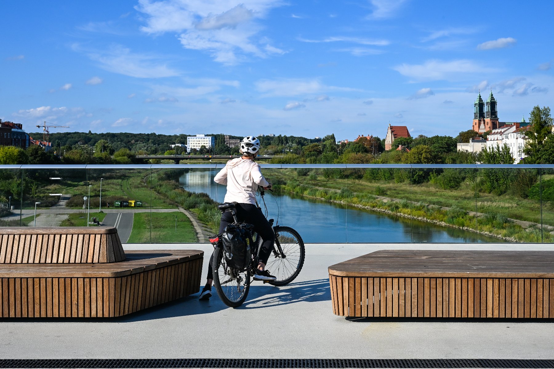 Kobieta w białym kasku i jasnej bluzie stoi z rowerem na nowoczesnej kładce, patrząc w stronę rzeki Warty. Rower wyposażony jest w czarne sakwy. Na pierwszym planie widoczne są fragmenty dwóch drewnianych ław o nieregularnym kształcie. W tle rozpościera się panorama Poznania z widocznymi wieżami katedry na Ostrowie Tumskim pod błękitnym niebem z białymi chmurami.
