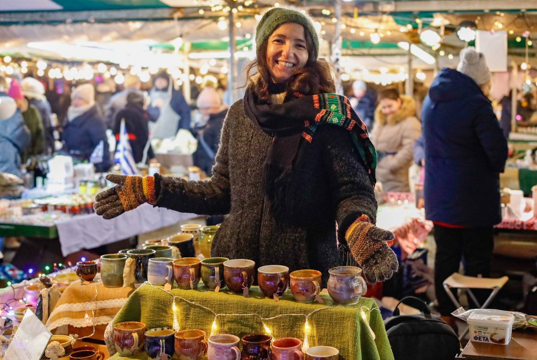 Kobieta w zimowym ubraniu, z wełnianą czapką i kolorowymi rękawiczkami, uśmiecha się szeroko, stojąc przy stoisku z ręcznie robioną ceramiką na świątecznym jarmarku. Na stole przed nią znajdują się kubki w różnych kolorach, oświetlone delikatnymi lampkami. W tle widoczni są odwiedzający w ciepłych ubraniach.