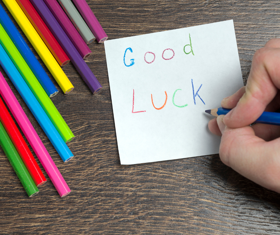 Kolorowe kredki leżące na drewnianym blacie obok kartki z napisem "Good Luck", który jest zapisywany niebieską kredką przez trzymaną w dłoni osobę.