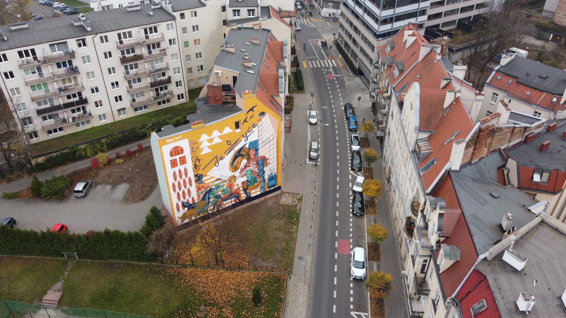 Kolorowy mural na ścianie budynku przedstawiający różnorodne elementy, w tym ludzi, ptaka i abstrakcyjne kształty. Na muralu znajdują się również napisy w innym języku. Po obu stronach ulicy zaparkowane samochody. Budynki po lewej stronie mają balkony, a budynki po prawej stronie mają dachy z czerwonych dachówek. Obrazek pokazuje mieszankę nowoczesnej i starszej architektury w środowisku miejskim.