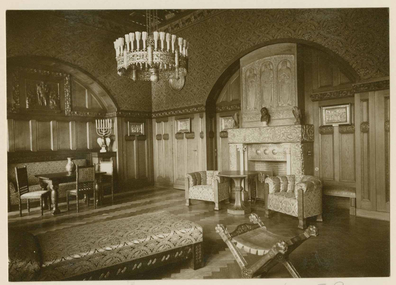 Komntaa zamkowa. Kominek w centrum. Obok stolik i dwa fotele. Duża ozdobna lampa i dywan. Drewniana boazeria.