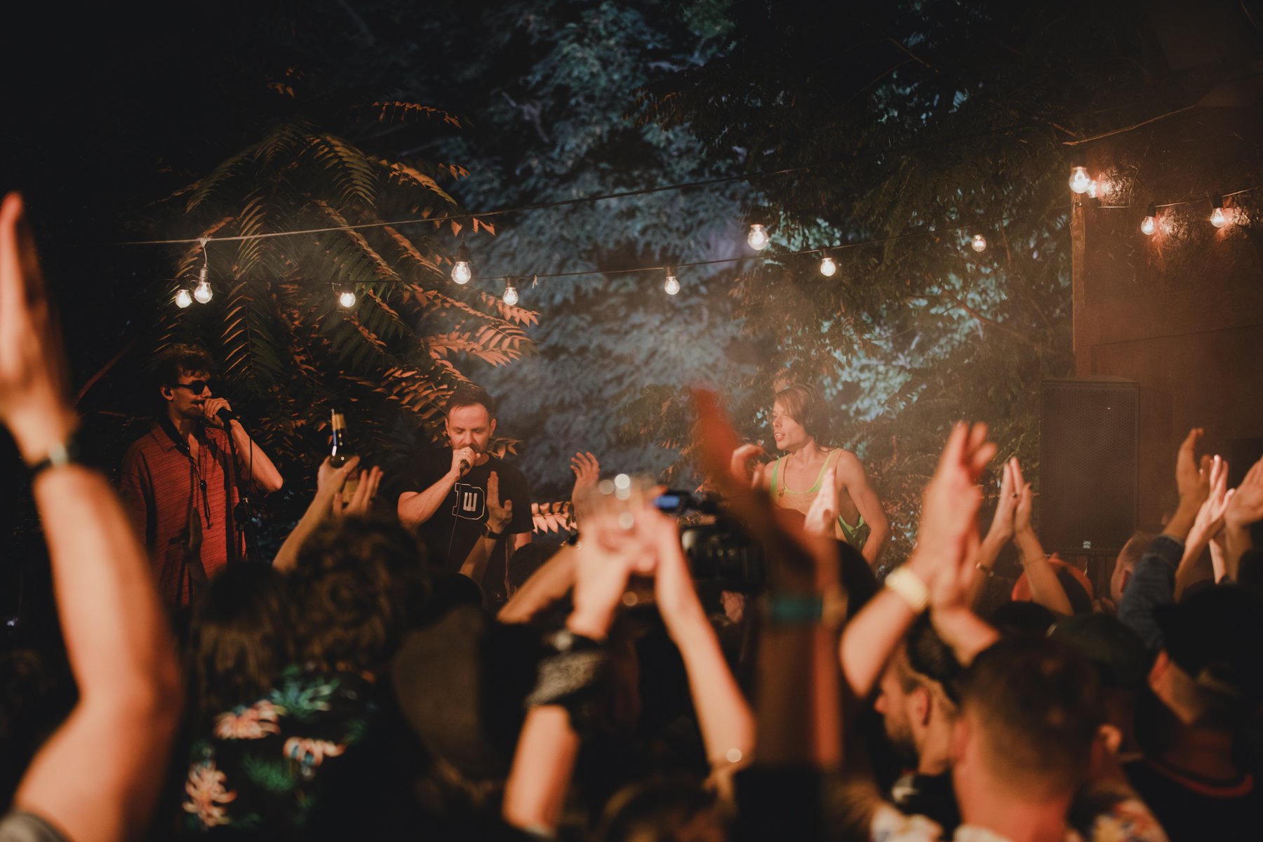 Koncert w pomieszczeniu oświetlonym wiszącymi żarówkami i czerwonym światłem. Na scenie trzech artystów śpiewa do mikrofonu. Na pierwszym planie widoczny tłum publiczności z uniesionymi rękami.