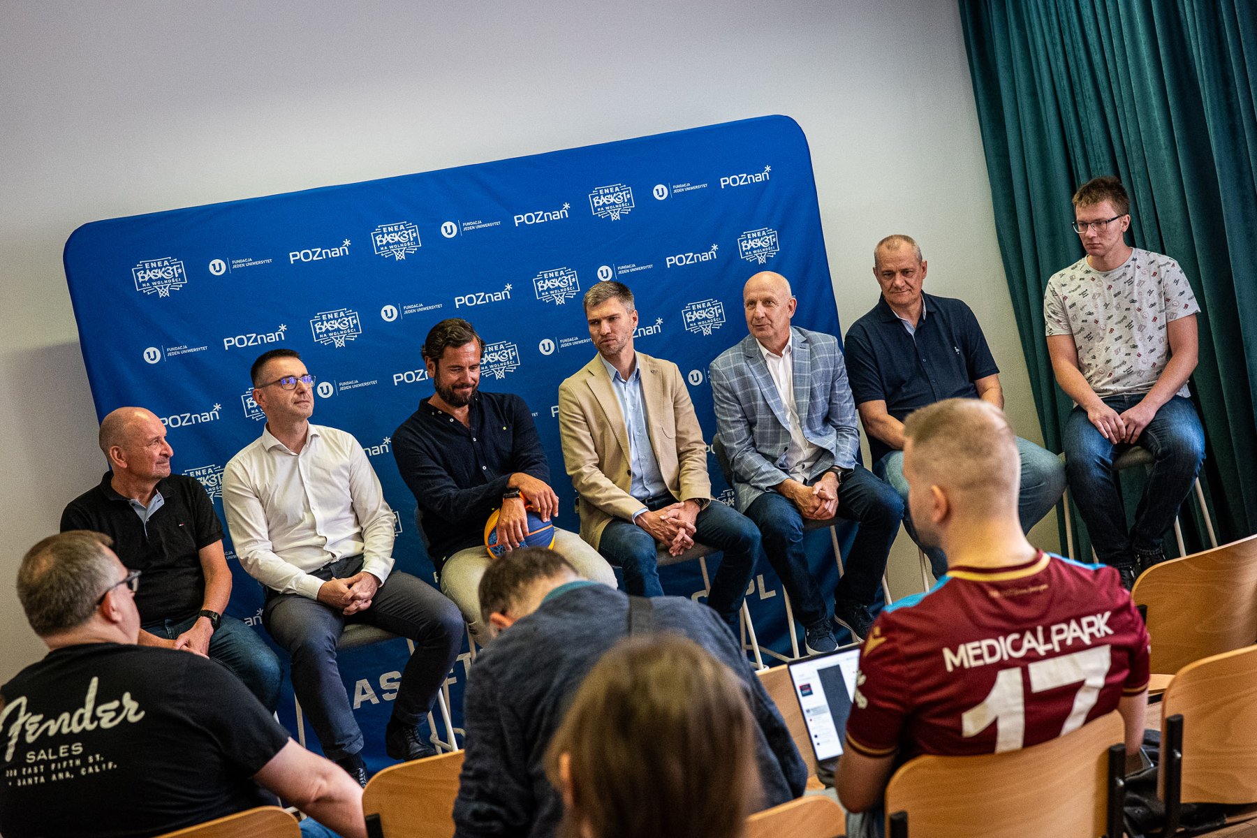 Konferencja prasowa przed rozpoczęciem Enea Basket na Wolności 2026, fot. Enea Basket na Wolności 2026 / Piotrek Przyborowski - na tle ścianki siedzi kilka osób, z których jedna osoba trzyma piłkę. Tyłem siedzi kilka osób, które się przysłuchuje