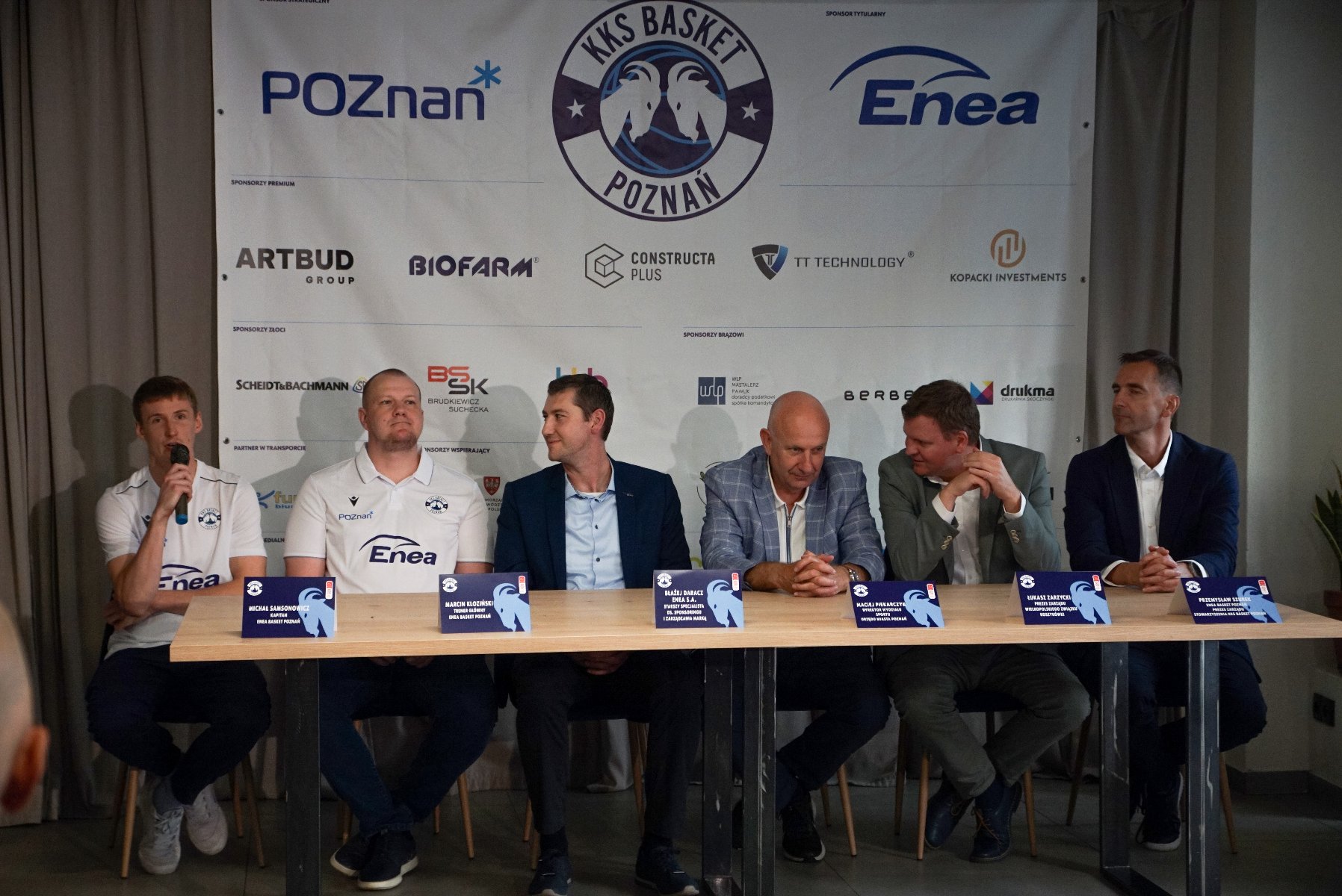 Konferencja prasowa przed sezonem Enea Basket Poznań, fot. Beata Brociek - za stołem siedzi 6 mężczyzn na tle klubowego baneru. Mężczyzna z lewej strony trzyma mikrofon
