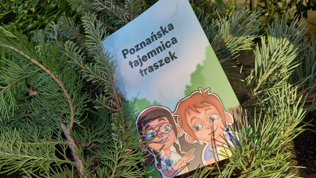 Książeczka dla dzieci zatytułowana "Poznańska tajemnica traszek" leży oparta o gałązki iglaków. Na okładce widnieje ilustracja dwójki uśmiechniętych dzieci - dziewczynki w okularach i chłopca - patrzących na małą traszkę trzymaną na dłoni.