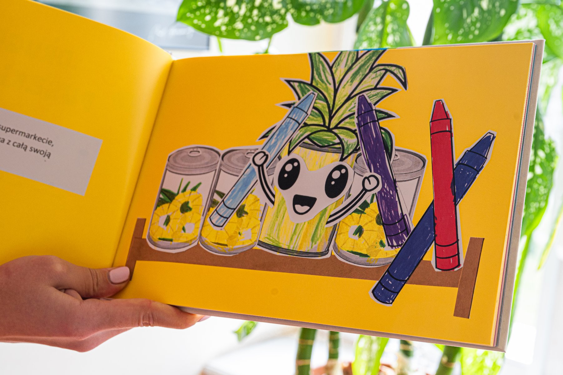 Książka dla dzieci z jasnym żółtym tłem. Na prawej stronie ilustracja uśmiechniętego ananas z dużymi, wyrazistymi oczami i szerokim uśmiechem. Ananas trzyma dwa kredki: niebieską i fioletową, a obok, na półce, opierają się dwie inne kredki: czerwona i niebieska. Na półce znajdują się również trzy puszki z plasterkami ananasa. Na lewej stronie jest mały blok tekstu w języku polskim: "supermarkecie, z całą swoją". Książka jest trzymana przez osobę z różowym lakierem do paznokci.