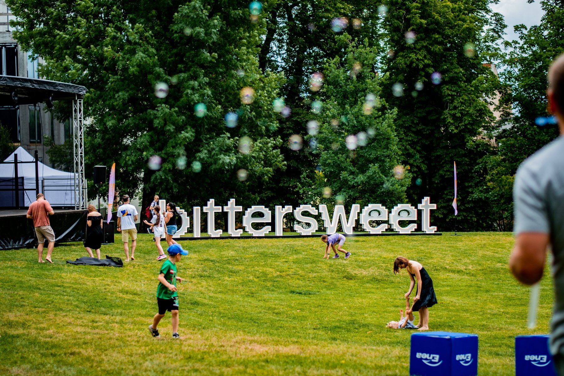 Ludzie bawią się na zielonej polanie. Na trawie znajdują się duże, białe litery układające się w napis "bittersweet". W tle widać scenę, drzewa i latające bańki mydlane.