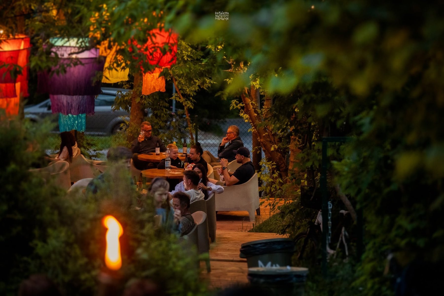 Ludzie siedzą w plenerowym ogródku restauracyjnym wśród kolorowych lampionów.