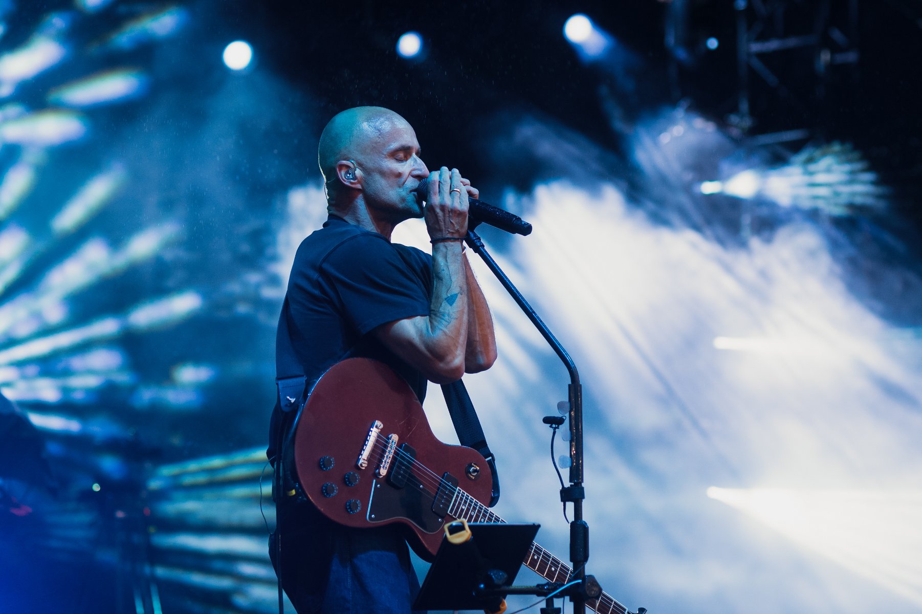 Łysy mężczyna przy mikrofonie, z gitarą na scenie.