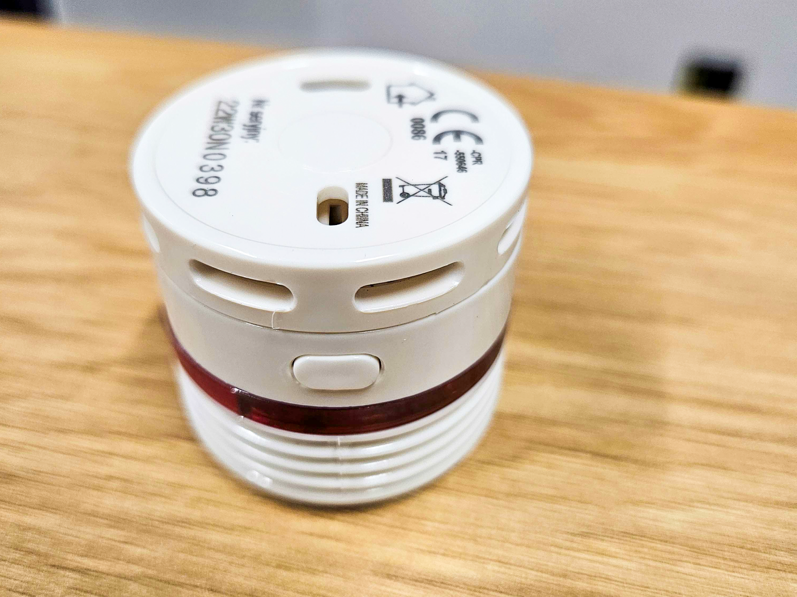 Mała, okrągła, biała czujka dymu lub ciepła, leżąca na drewnianym blacie. Urządzenie ma czerwoną opaskę świetlną dookoła i widoczne otwory wentylacyjne oraz przycisk.