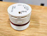 Mała, okrągła, biała czujka dymu lub ciepła, leżąca na drewnianym blacie. Urządzenie ma czerwoną opaskę świetlną dookoła i widoczne otwory wentylacyjne oraz przycisk.