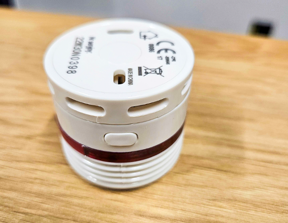 Mała, okrągła, biała czujka dymu lub ciepła, leżąca na drewnianym blacie. Urządzenie ma czerwoną opaskę świetlną dookoła i widoczne otwory wentylacyjne oraz przycisk.