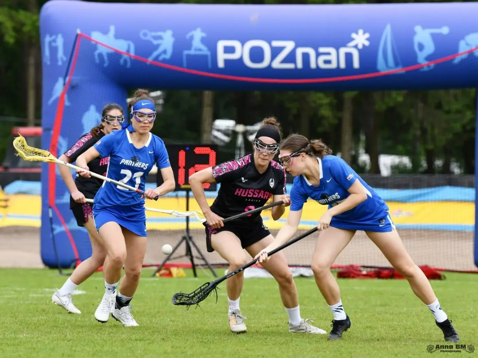 Mecz lacrosse na świeżym powietrzu. Na trawiastym boisku cztery zawodniczki z kijami, ubrane w sportowe stroje, walczą o piłkę. W tle dmuchana, niebieska brama z napisem "POZnań". Na dole, w prawym rogu, widnieje podpis fotografa: "Anna BM FOTOGRAFIA".