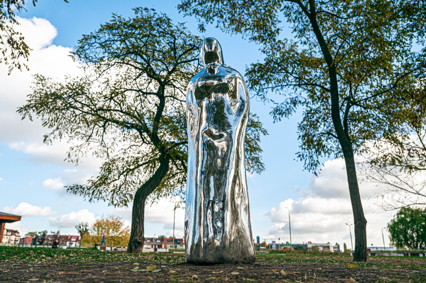 Metaliczna, błyszcząca statua w parku. Przedstawia ludzką postać w płaszczu, o gładkiej, lustrzanej powierzchni, która odbija otaczającą przestrzeń.